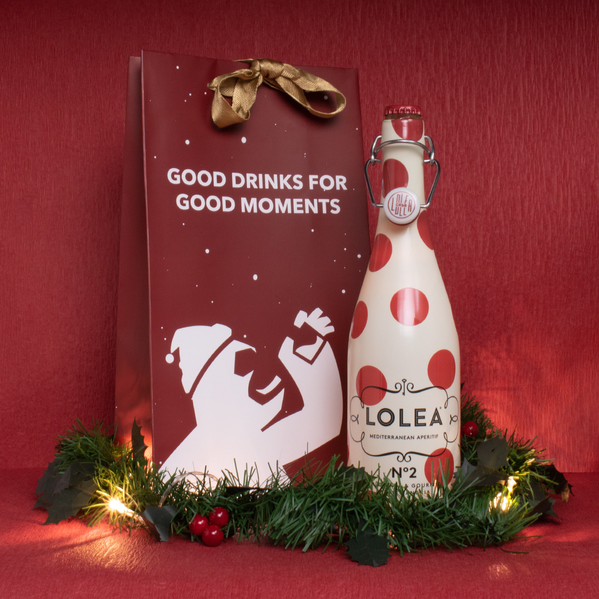 Christmas edition carrier bag in maroon with a Santa-hat gorilla, beside a bottle of Lolea sangria on pine and fairy lights